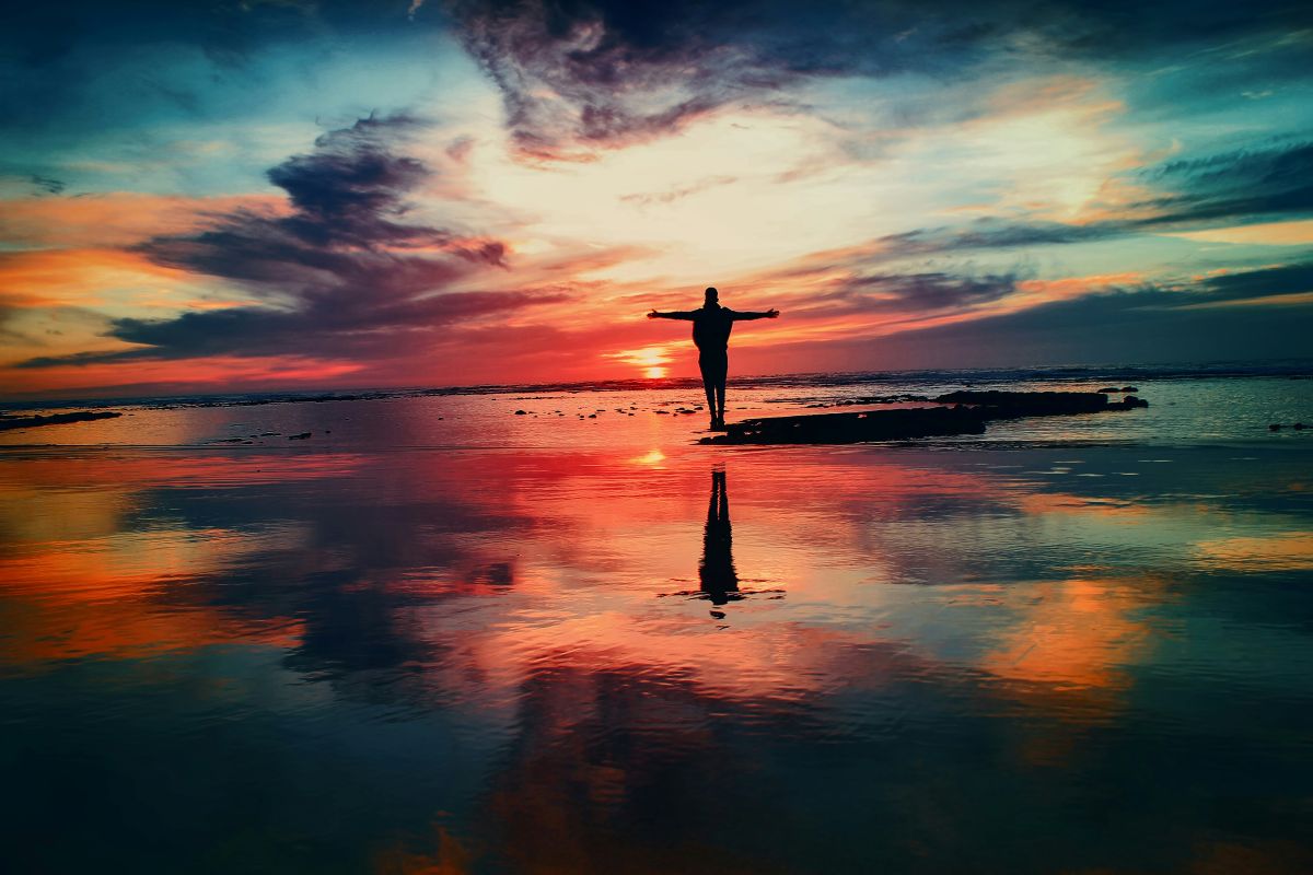 Man standing with outstretched arms. The background sky is colorful and changing shape.