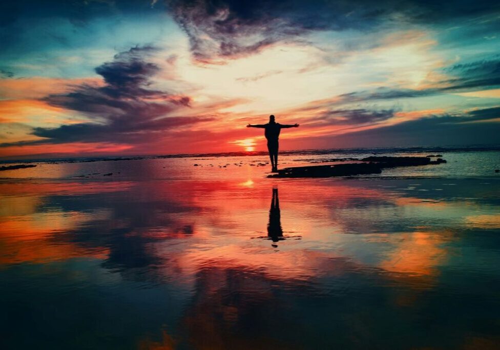 Man standing with outstretched arms. The background sky is colorful and changing shape.